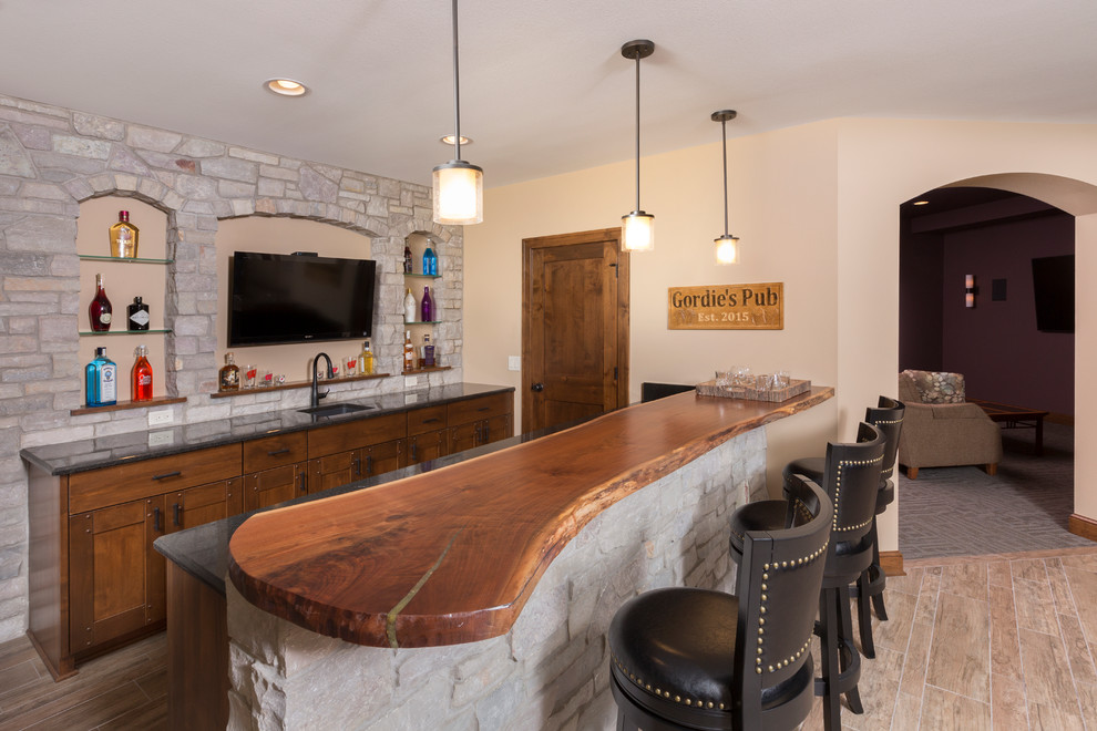 Large country galley seated home bar in Milwaukee with an undermount sink, medium wood cabinets, wood benchtops, grey splashback, stone tile splashback, ceramic floors, brown benchtop, shaker cabinets and beige floor.