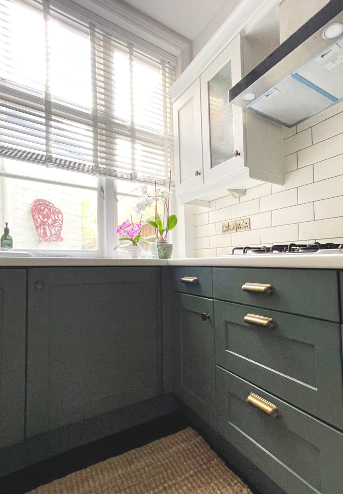 Photo of a medium sized traditional cream and black l-shaped kitchen/diner in London with a belfast sink, shaker cabinets, green cabinets, laminate countertops, beige splashback, ceramic splashback, stainless steel appliances, painted wood flooring, no island, black floors and a wallpapered ceiling.