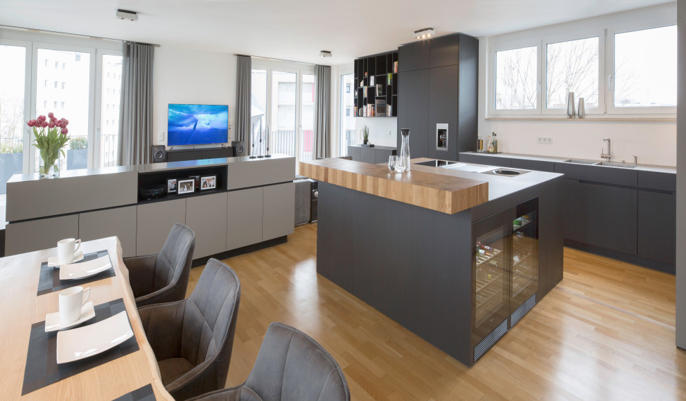 Einzeilige Moderne Wohnküche mit integriertem Waschbecken, flächenbündigen Schrankfronten, schwarzen Schränken, Edelstahl-Arbeitsplatte, Küchengeräten aus Edelstahl, hellem Holzboden und Kücheninsel in München