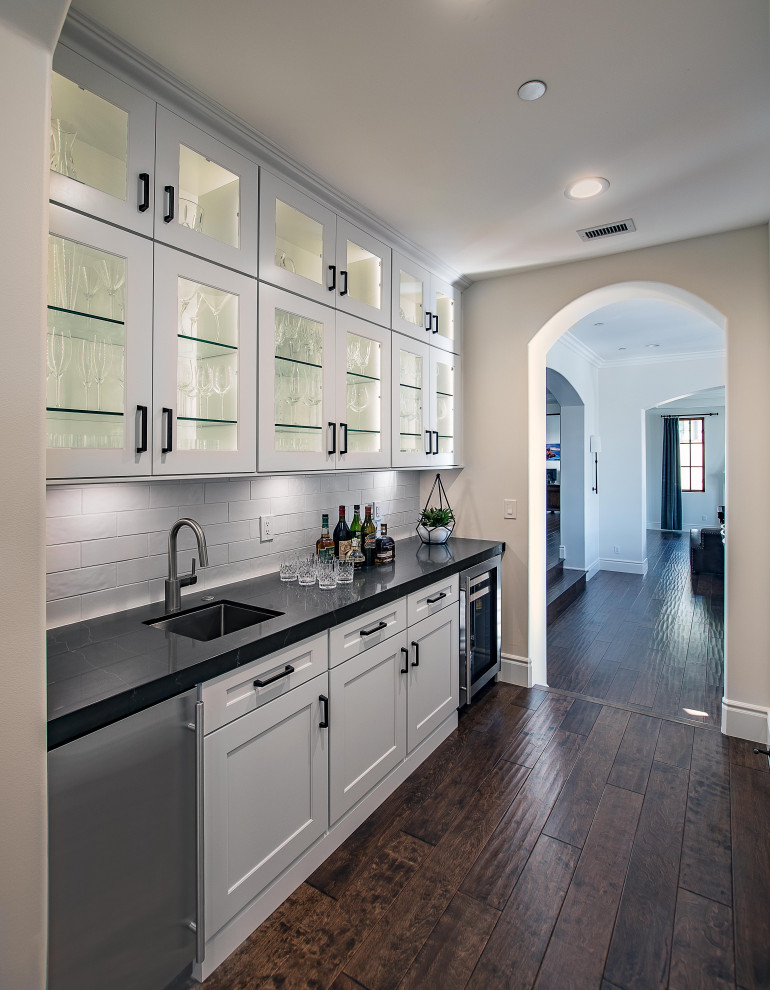 This is an example of a transitional single-wall kitchen pantry in Orange County with an undermount sink, glass-front cabinets, white cabinets, white splashback, subway tile splashback, dark hardwood floors, no island, brown floor and black benchtop.