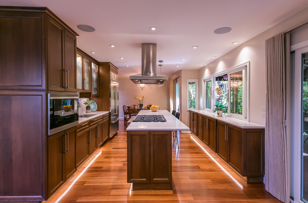This is an example of a large contemporary galley eat-in kitchen in San Francisco with an undermount sink, recessed-panel cabinets, dark wood cabinets, quartz benchtops, white splashback, stone slab splashback, stainless steel appliances, with island, brown floor, medium hardwood floors and white benchtop.
