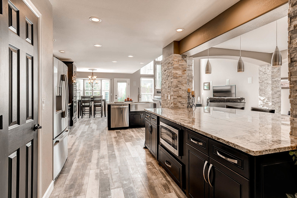 Photo of a large contemporary l-shaped open plan kitchen in Denver with raised-panel cabinets, dark wood cabinets, granite benchtops, grey splashback, stone tile splashback, stainless steel appliances, grey floor, a farmhouse sink, laminate floors, multiple islands and multi-coloured benchtop.
