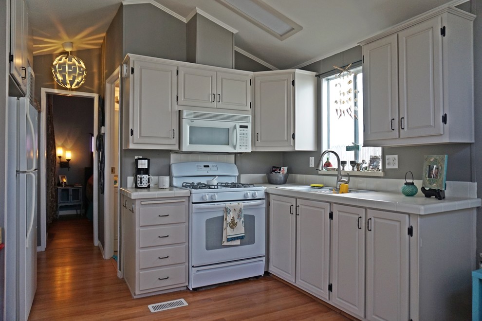 Photo of a small contemporary l-shaped eat-in kitchen in San Diego with an undermount sink, raised-panel cabinets, white cabinets, tile benchtops, white splashback, porcelain splashback, white appliances, medium hardwood floors and no island.