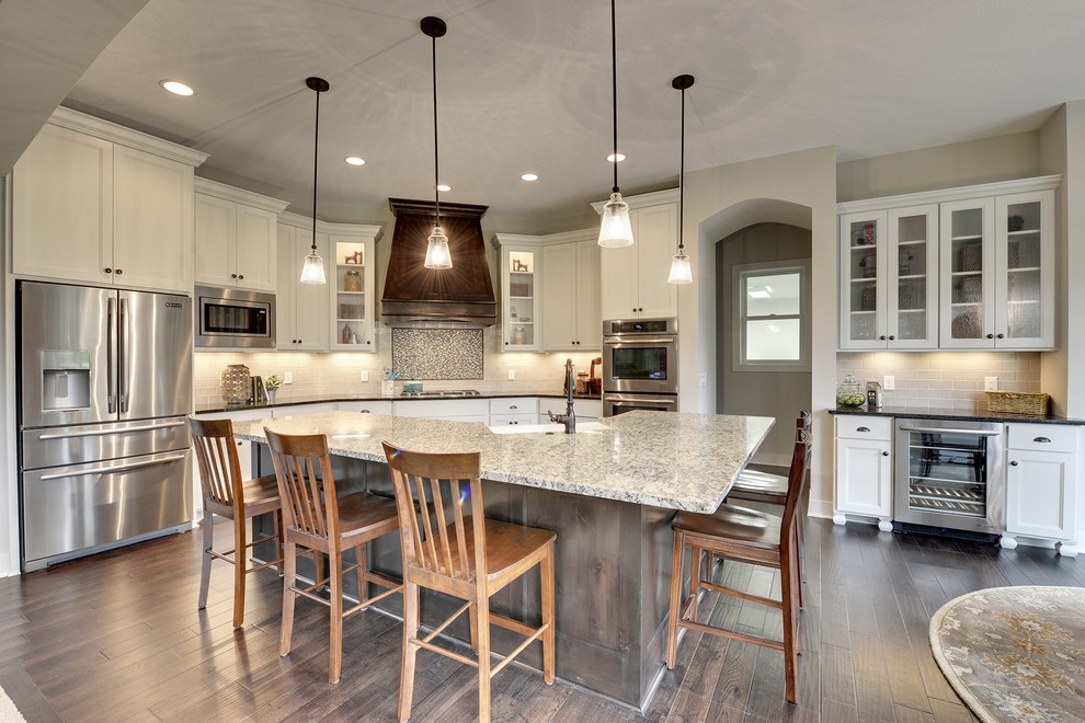 This is an example of a large transitional l-shaped open plan kitchen in Minneapolis with recessed-panel cabinets, white cabinets, with island, a farmhouse sink, grey splashback, glass tile splashback, stainless steel appliances and dark hardwood floors.