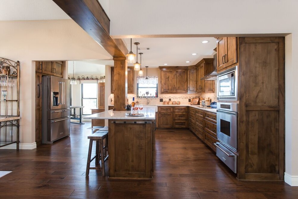 This is an example of a large transitional l-shaped eat-in kitchen in Dallas with a farmhouse sink, recessed-panel cabinets, medium wood cabinets, multi-coloured splashback, with island, brown floor, quartzite benchtops, travertine splashback, stainless steel appliances, medium hardwood floors and white benchtop.