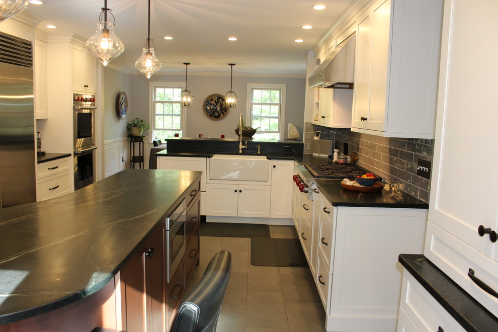 Photo of a large traditional u-shaped eat-in kitchen in Boston with a farmhouse sink, recessed-panel cabinets, white cabinets, granite benchtops, grey splashback, subway tile splashback, stainless steel appliances, porcelain floors, with island, grey floor and black benchtop.