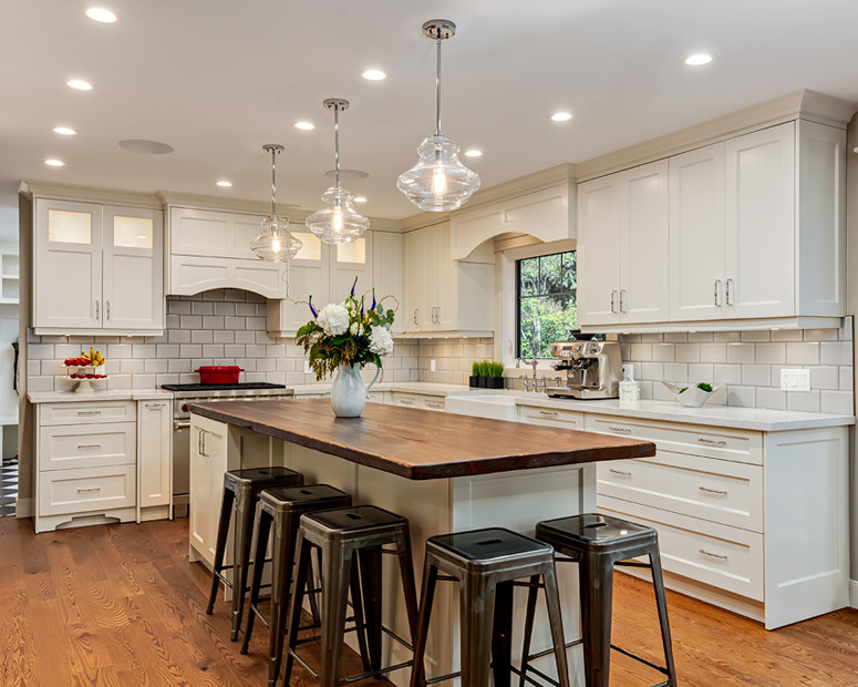 Example of a mid-sized transitional l-shaped light wood floor and beige floor enclosed kitchen design in Calgary with a farmhouse sink, recessed-panel cabinets, white cabinets, quartz countertops, white backsplash, porcelain backsplash, stainless steel appliances and an island