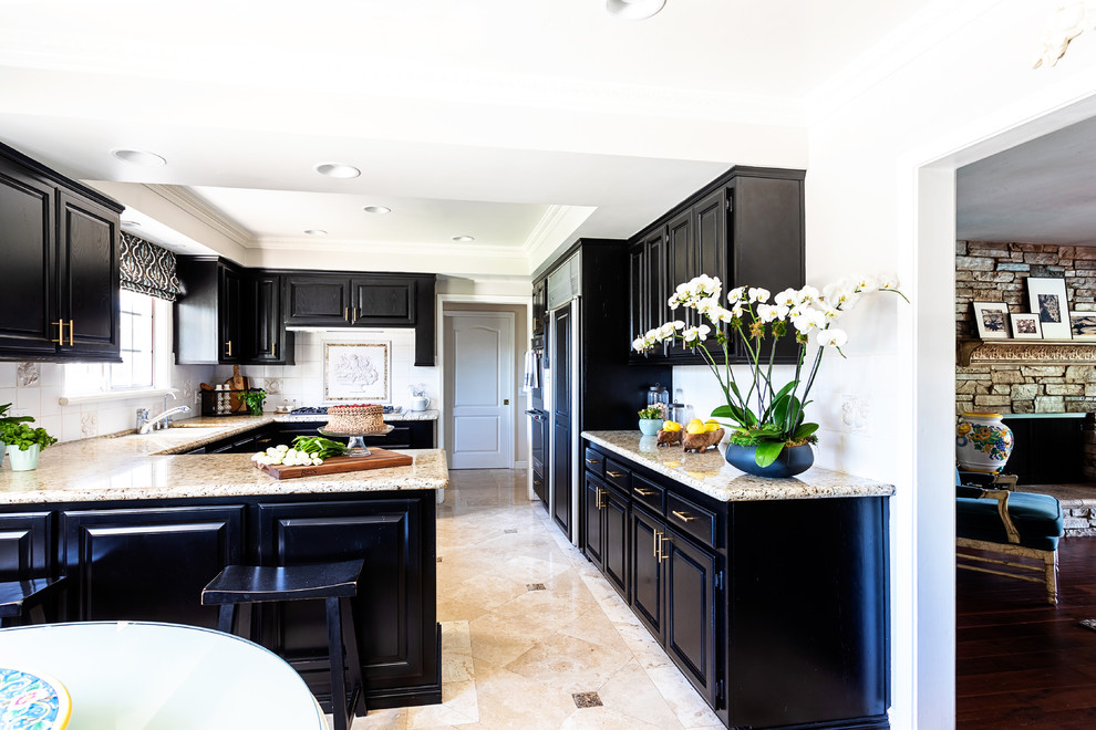 Mid-sized traditional u-shaped eat-in kitchen in Los Angeles with an undermount sink, raised-panel cabinets, black cabinets, granite benchtops, white splashback, ceramic splashback, stainless steel appliances, travertine floors, beige floor and beige benchtop.