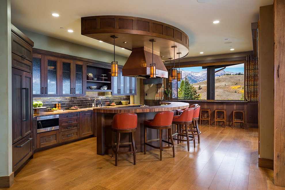 Mountain style galley medium tone wood floor open concept kitchen photo in Denver with glass-front cabinets, dark wood cabinets, wood countertops, brown backsplash, subway tile backsplash, paneled appliances and an island