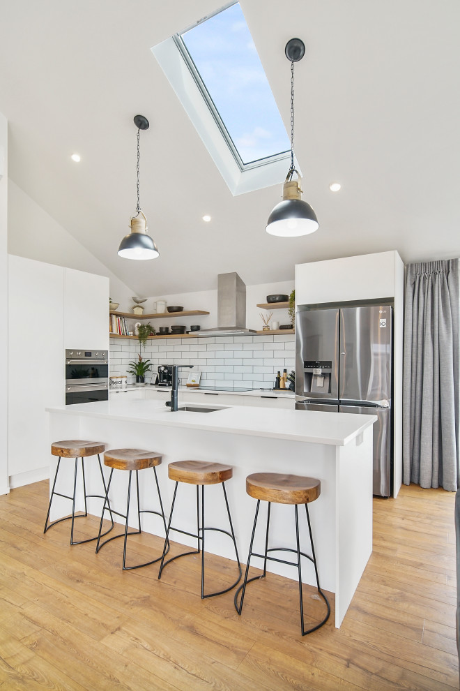 Beach style l-shaped kitchen in Auckland with an undermount sink, flat-panel cabinets, white cabinets, white splashback, subway tile splashback, stainless steel appliances, medium hardwood floors, with island, brown floor, white benchtop and vaulted.
