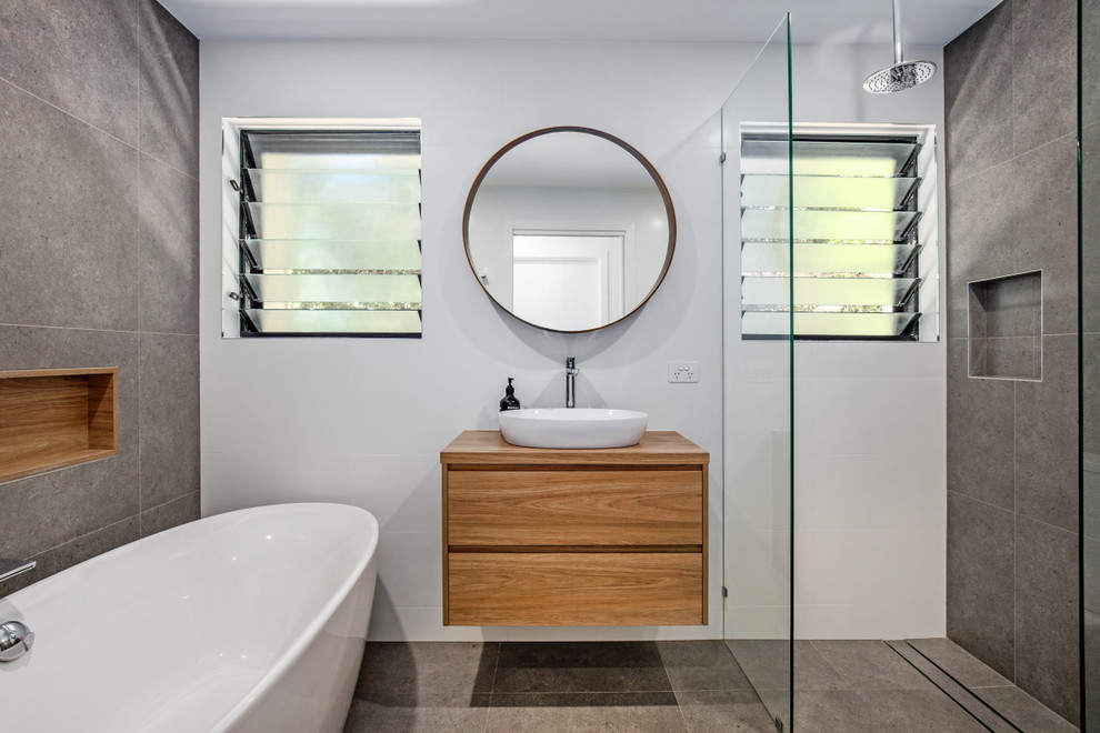 This is an example of a mid-sized beach style kids bathroom in Sunshine Coast with furniture-like cabinets, light wood cabinets, a freestanding tub, an open shower, gray tile, cement tile, grey walls, porcelain floors, a vessel sink, wood benchtops, grey floor and an open shower.