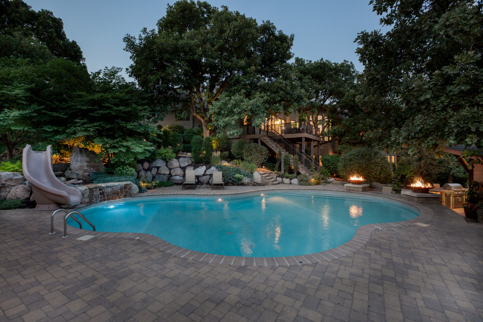 This is an example of a traditional backyard custom-shaped pool in Omaha with a water slide and concrete pavers.