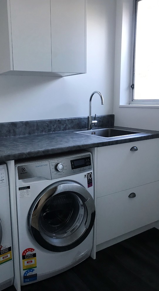 Photo of a small contemporary l-shaped dedicated laundry room in Christchurch with a single-bowl sink, flat-panel cabinets, white cabinets, laminate benchtops, white walls, vinyl floors, a side-by-side washer and dryer, grey floor and grey benchtop.