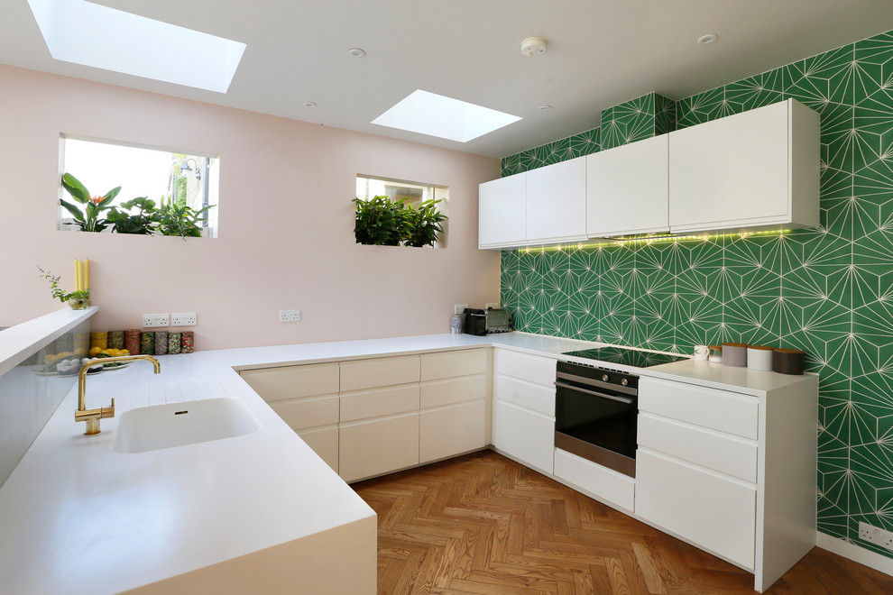This is an example of a contemporary u-shaped kitchen in Other with an integrated sink, flat-panel cabinets, white cabinets, green splashback, stainless steel appliances, medium hardwood floors, a peninsula, brown floor and white benchtop.