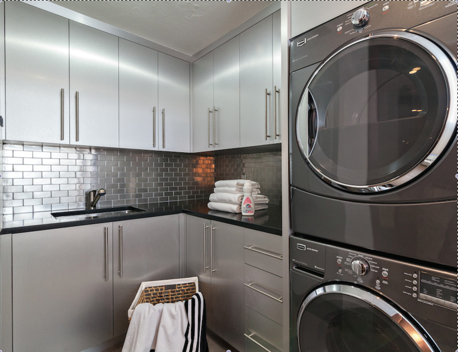 Inspiration for a mid-sized modern l-shaped dedicated laundry room in Miami with an undermount sink, flat-panel cabinets, grey cabinets, granite benchtops, grey walls, travertine floors, a stacked washer and dryer, beige floor and black benchtop.