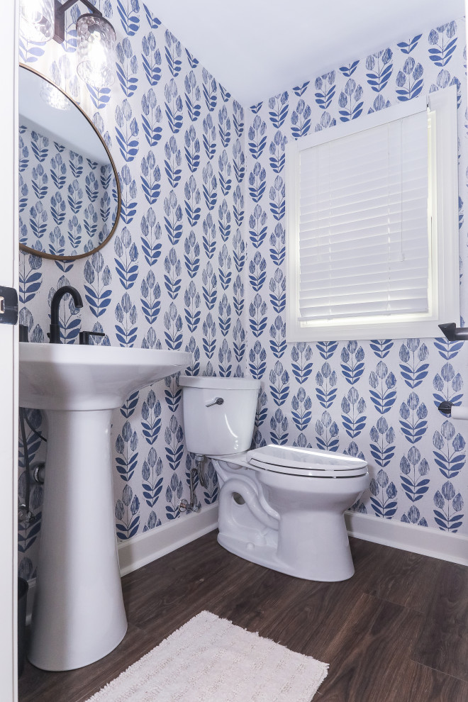 Small transitional 3/4 bathroom with blue walls, a pedestal sink, a single vanity, a freestanding vanity, laminate floors, brown floor, an enclosed toilet and wallpaper.