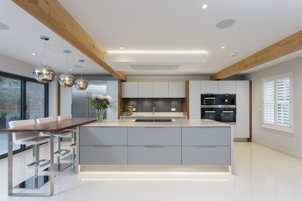 Photo of a contemporary galley kitchen in Sussex with an undermount sink, flat-panel cabinets, grey cabinets, grey splashback, glass sheet splashback, black appliances, with island, white floor and white benchtop.