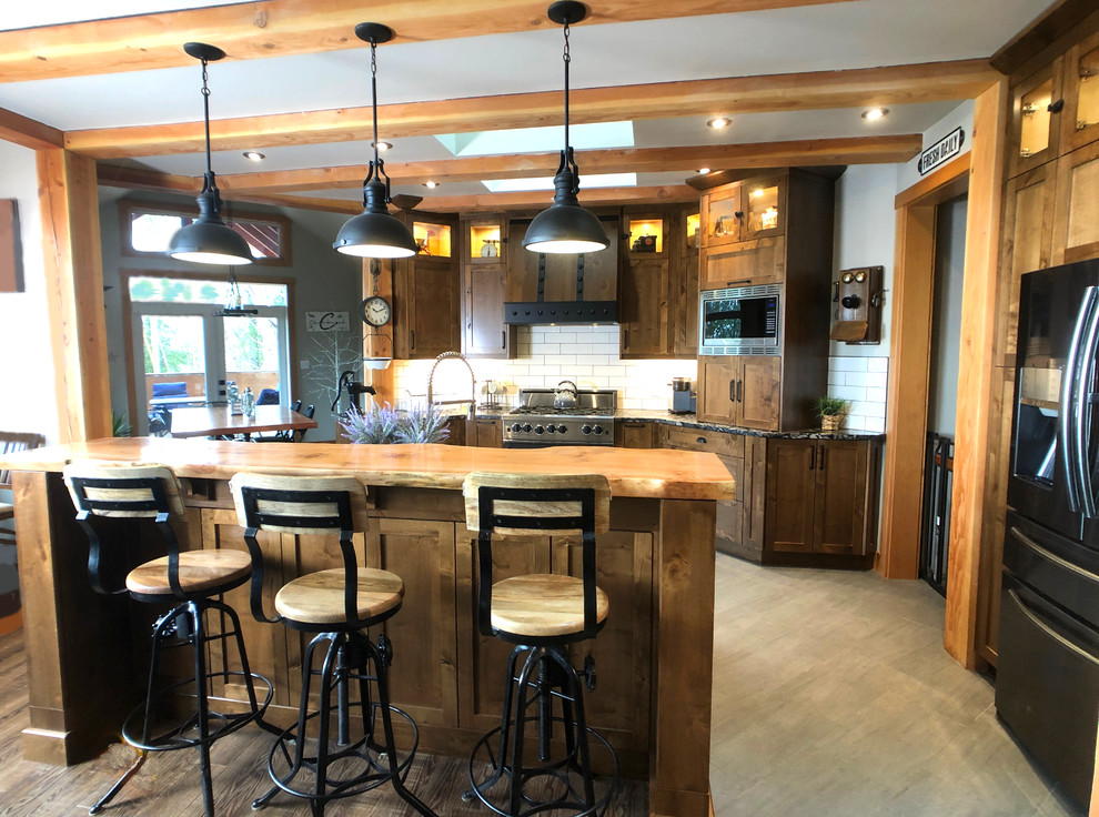 Photo of a large industrial l-shaped eat-in kitchen in Vancouver with a farmhouse sink, shaker cabinets, medium wood cabinets, white splashback, subway tile splashback, stainless steel appliances, light hardwood floors, with island, brown floor and black benchtop.