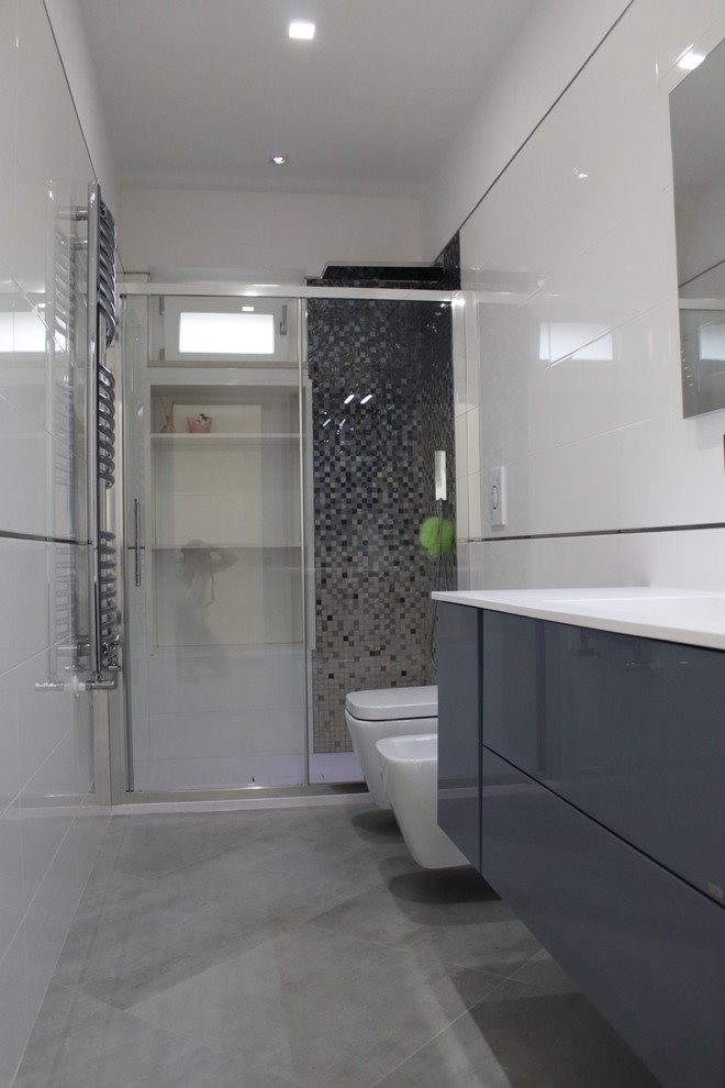 Bild på ett mellanstort funkis vit vitt badrum med dusch, med släta luckor, grå skåp, en kantlös dusch, en toalettstol med separat cisternkåpa, vit kakel, porslinskakel, vita väggar, klinkergolv i porslin, ett integrerad handfat, bänkskiva i akrylsten, grått golv och dusch med skjutdörr
