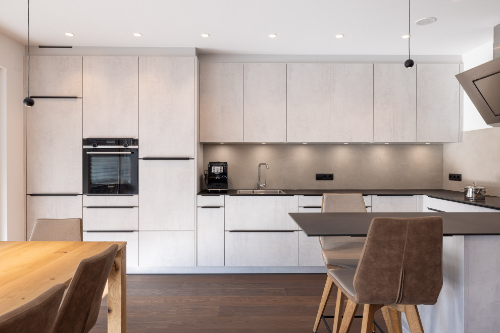 Mid-sized contemporary u-shaped open plan kitchen in Other with a double-bowl sink, flat-panel cabinets, grey cabinets, solid surface benchtops, grey splashback, ceramic splashback, black appliances, dark hardwood floors, a peninsula, brown floor and black benchtop.