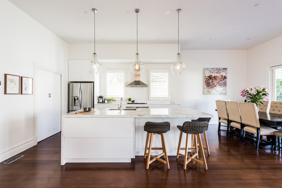 Photo of a mid-sized contemporary single-wall open plan kitchen in Melbourne with an undermount sink, flat-panel cabinets, white cabinets, marble benchtops, white splashback, subway tile splashback, stainless steel appliances, dark hardwood floors, with island and brown floor.