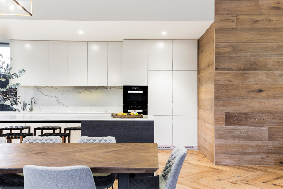 This is an example of a modern single-wall eat-in kitchen in Melbourne with an undermount sink, flat-panel cabinets, white cabinets, quartz benchtops, multi-coloured splashback, stone slab splashback, black appliances, light hardwood floors, with island and brown floor.