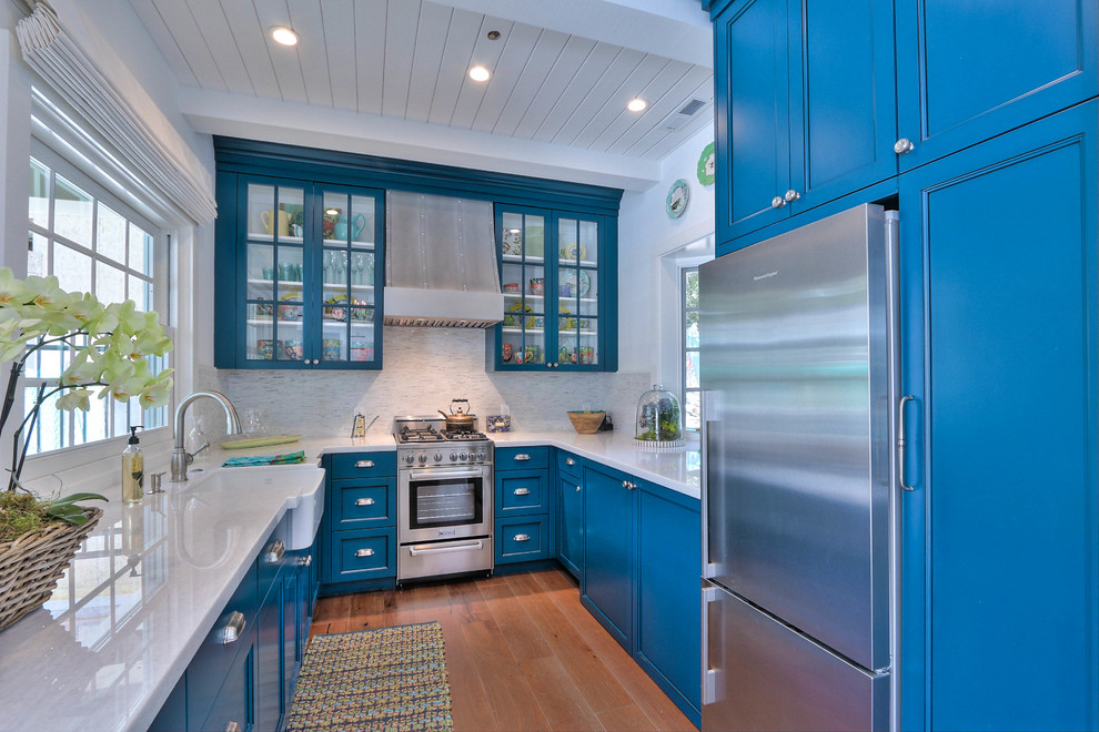 This is an example of a small beach style u-shaped separate kitchen in San Francisco with a farmhouse sink, blue cabinets, white splashback, stainless steel appliances, medium hardwood floors, glass-front cabinets and matchstick tile splashback.