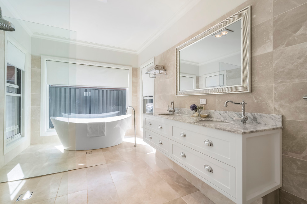 This is an example of a large contemporary master bathroom in Orange County with flat-panel cabinets, white cabinets, a freestanding tub, an open shower, a one-piece toilet, beige tile, stone tile, beige walls, slate floors, a drop-in sink, marble benchtops, beige floor, an open shower and white benchtops.