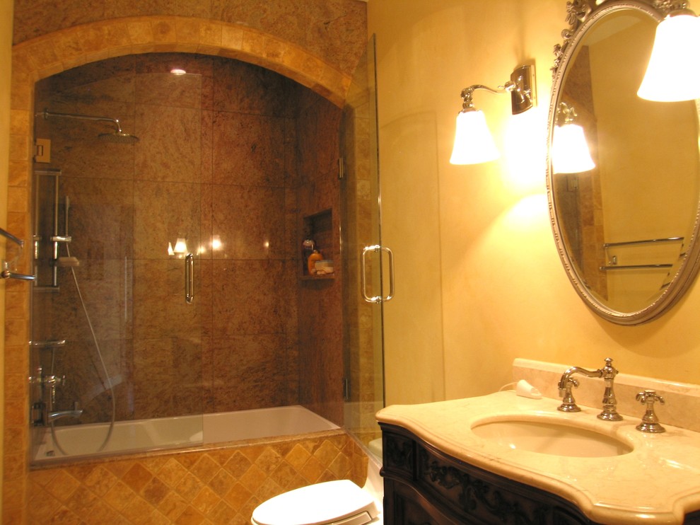 Photo of a small mediterranean master bathroom in San Diego with furniture-like cabinets, dark wood cabinets, an undermount tub, a one-piece toilet, brown tile, stone tile, beige walls, marble floors, an undermount sink, marble benchtops, beige floor and a hinged shower door.