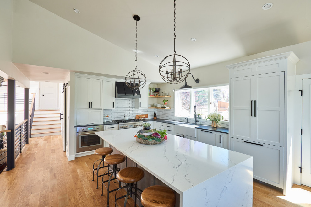This is an example of a large country l-shaped open plan kitchen in Denver with a farmhouse sink, shaker cabinets, white cabinets, quartz benchtops, white splashback, porcelain splashback, stainless steel appliances, medium hardwood floors, with island, brown floor and white benchtop.
