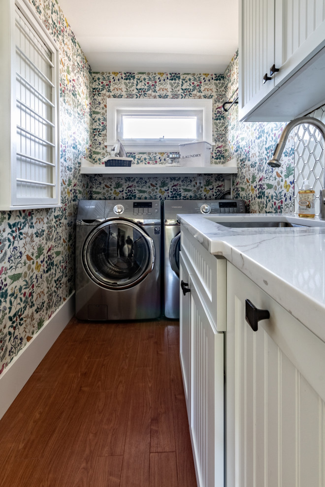 Photo of a small arts and crafts l-shaped dedicated laundry room in Vancouver with an undermount sink, white cabinets, quartz benchtops, white splashback, mosaic tile splashback, multi-coloured walls, laminate floors, a side-by-side washer and dryer, white benchtop, wallpaper, shaker cabinets and brown floor.