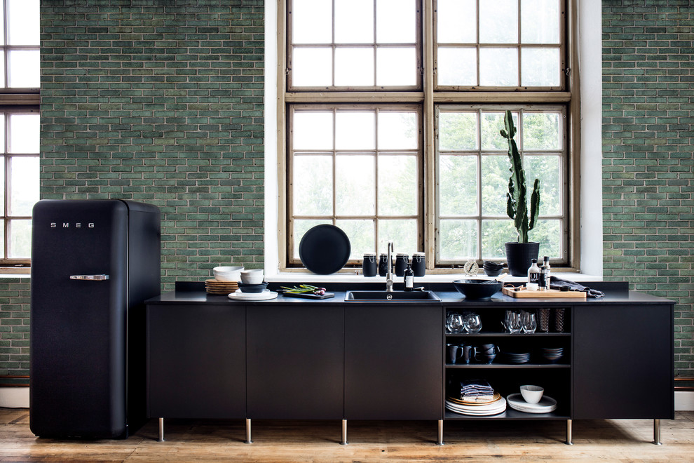 This is an example of a mid-sized industrial single-wall open plan kitchen in Gothenburg with a single-bowl sink, flat-panel cabinets, black cabinets, laminate benchtops, black appliances, light hardwood floors, no island, beige floor and black benchtop.