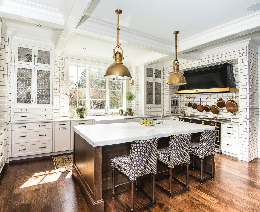 Photo of a country u-shaped open plan kitchen in Chicago with a farmhouse sink, glass-front cabinets, white cabinets, marble benchtops, white splashback, stone tile splashback, black appliances, medium hardwood floors, with island and brown floor.