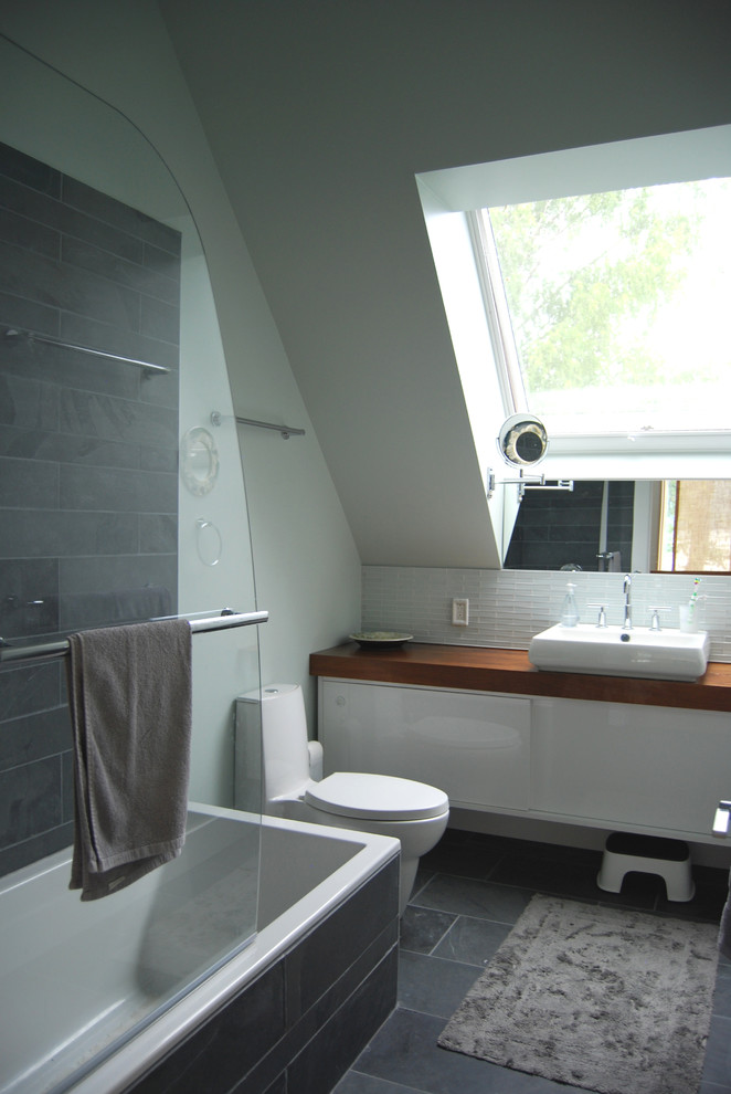 Photo of a modern kids bathroom in New York with flat-panel cabinets, white cabinets, a drop-in tub, a shower/bathtub combo, a one-piece toilet, gray tile, stone tile, green walls, slate floors, a vessel sink and wood benchtops.