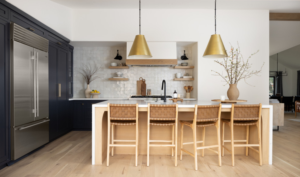 Example of a transitional l-shaped light wood floor and beige floor kitchen design in Orange County with an undermount sink, shaker cabinets, black cabinets, white backsplash, stainless steel appliances, an island and white countertops