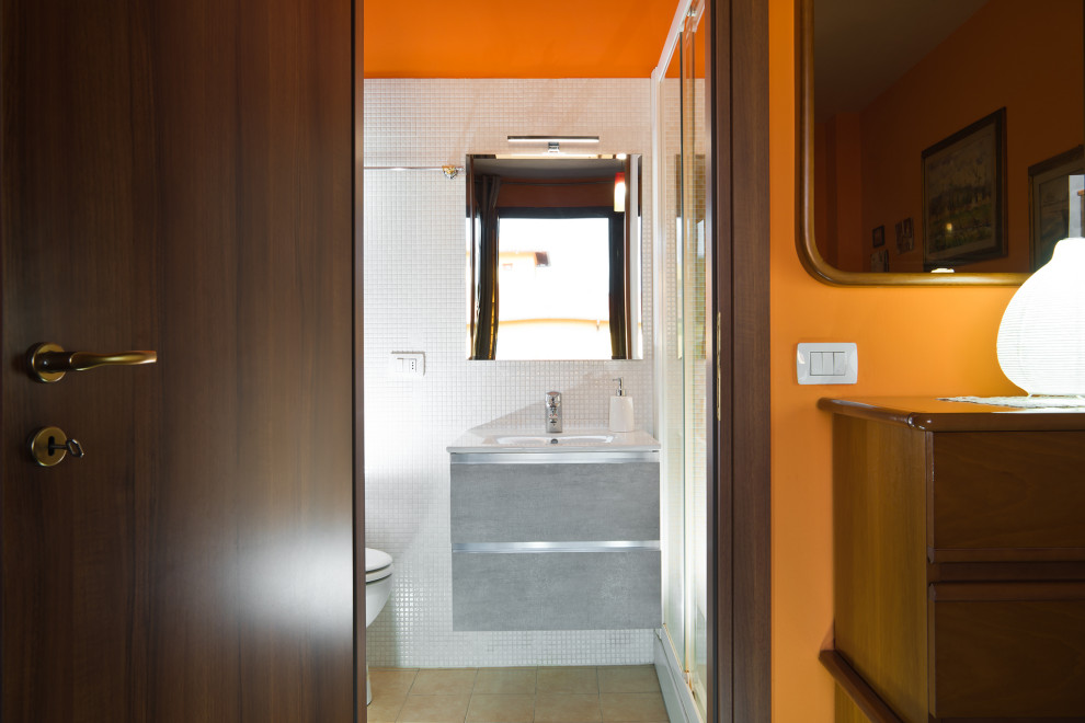 Photo of a small contemporary powder room in Florence with flat-panel cabinets, grey cabinets, a one-piece toilet, white tile, mosaic tile, orange walls, ceramic floors, an integrated sink, glass benchtops, beige floor, grey benchtops and a floating vanity.