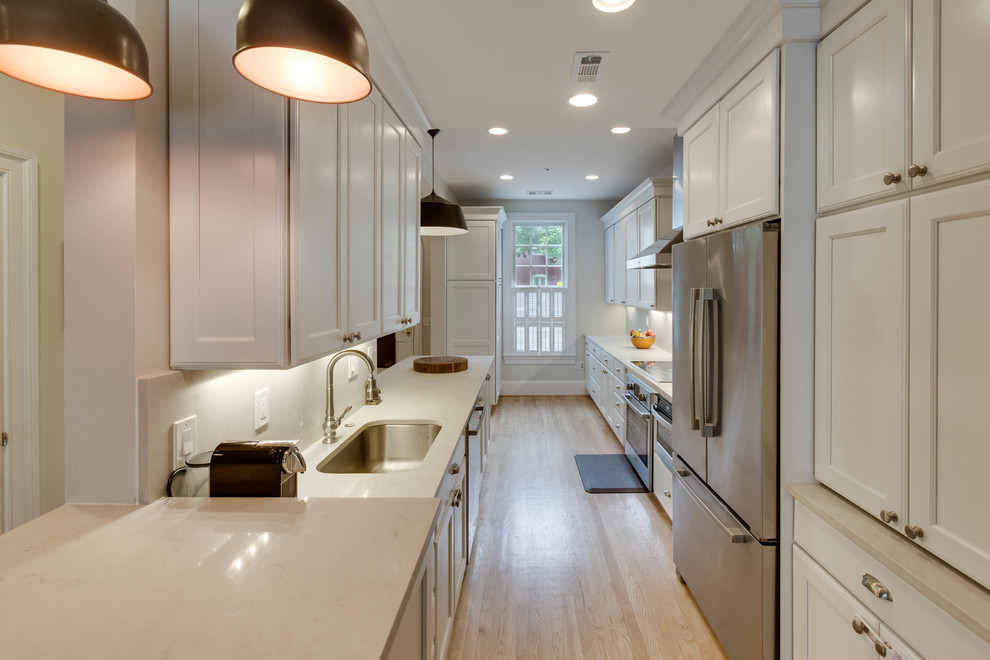 Photo of a mid-sized transitional galley kitchen in DC Metro with an undermount sink, recessed-panel cabinets, white cabinets, quartz benchtops, stainless steel appliances, light hardwood floors and a peninsula.
