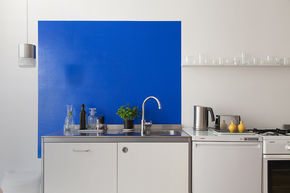 This is an example of a small contemporary single-wall open plan kitchen in Berlin with an integrated sink, flat-panel cabinets, white cabinets, stainless steel benchtops, blue splashback, limestone splashback, white appliances, light hardwood floors, no island and brown floor.