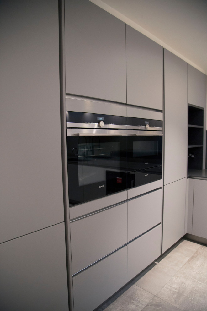 Photo of a contemporary l-shaped eat-in kitchen in London with a single-bowl sink, flat-panel cabinets, grey cabinets, quartzite benchtops, black splashback, mirror splashback, black appliances, porcelain floors, with island, grey floor and grey benchtop.