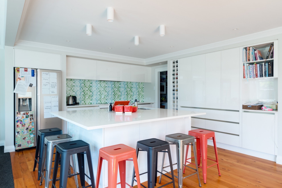 Large contemporary u-shaped kitchen in Sydney with a drop-in sink, multi-coloured splashback, glass sheet splashback, medium hardwood floors, with island, brown floor and white benchtop.
