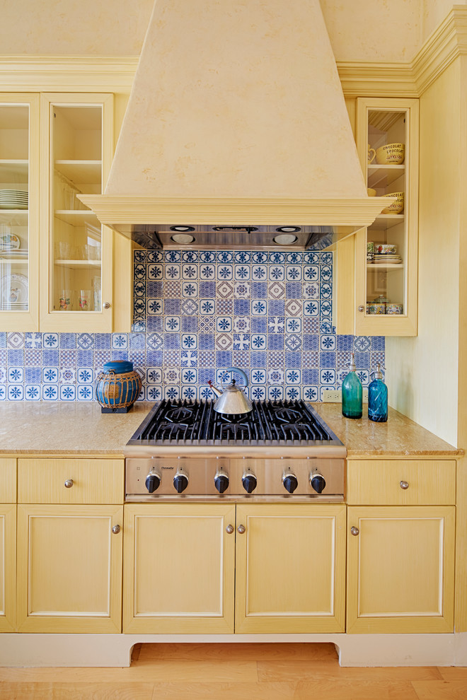 Design ideas for a mediterranean kitchen in San Francisco with glass-front cabinets, yellow cabinets, granite benchtops, stainless steel appliances, light hardwood floors and blue splashback.