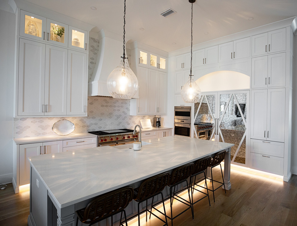 Inspiration for a large transitional l-shaped eat-in kitchen in Miami with a farmhouse sink, shaker cabinets, white cabinets, quartzite benchtops, white splashback, ceramic splashback, stainless steel appliances, medium hardwood floors, with island, beige floor, white benchtop and recessed.