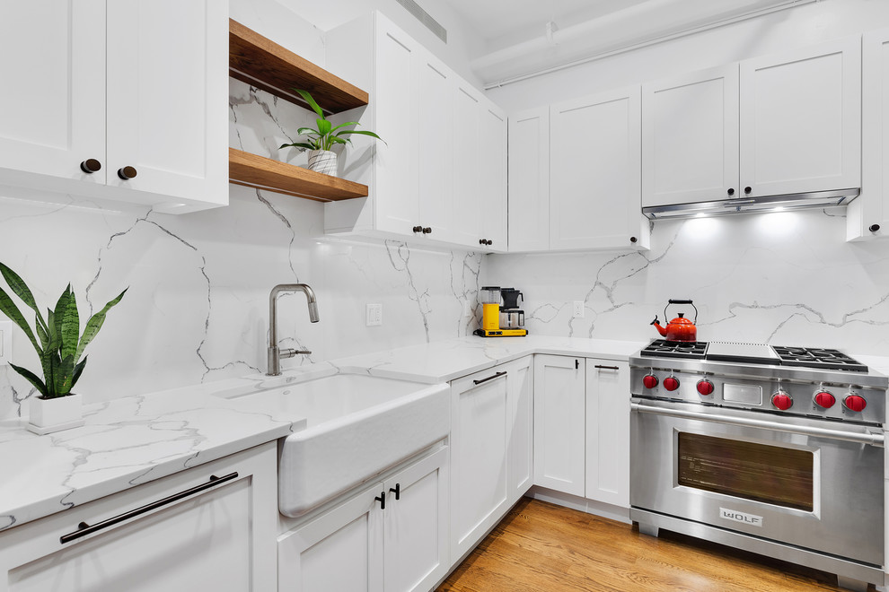 Photo of a transitional kitchen in New York with a farmhouse sink, shaker cabinets, white cabinets, quartz benchtops, white splashback, light hardwood floors and white benchtop.