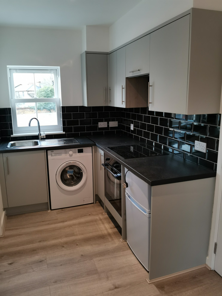 This is an example of a small modern grey and black l-shaped open plan kitchen in Cambridgeshire with a double-bowl sink, flat-panel cabinets, grey cabinets, laminate countertops, black splashback, ceramic splashback, white appliances, laminate floors, grey floors, black worktops and a drop ceiling.