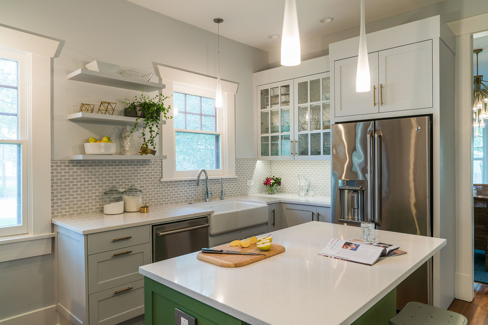 This is an example of a mid-sized transitional l-shaped kitchen in Austin with a farmhouse sink, shaker cabinets, grey splashback, glass tile splashback, stainless steel appliances, medium hardwood floors, with island, brown floor and grey cabinets.
