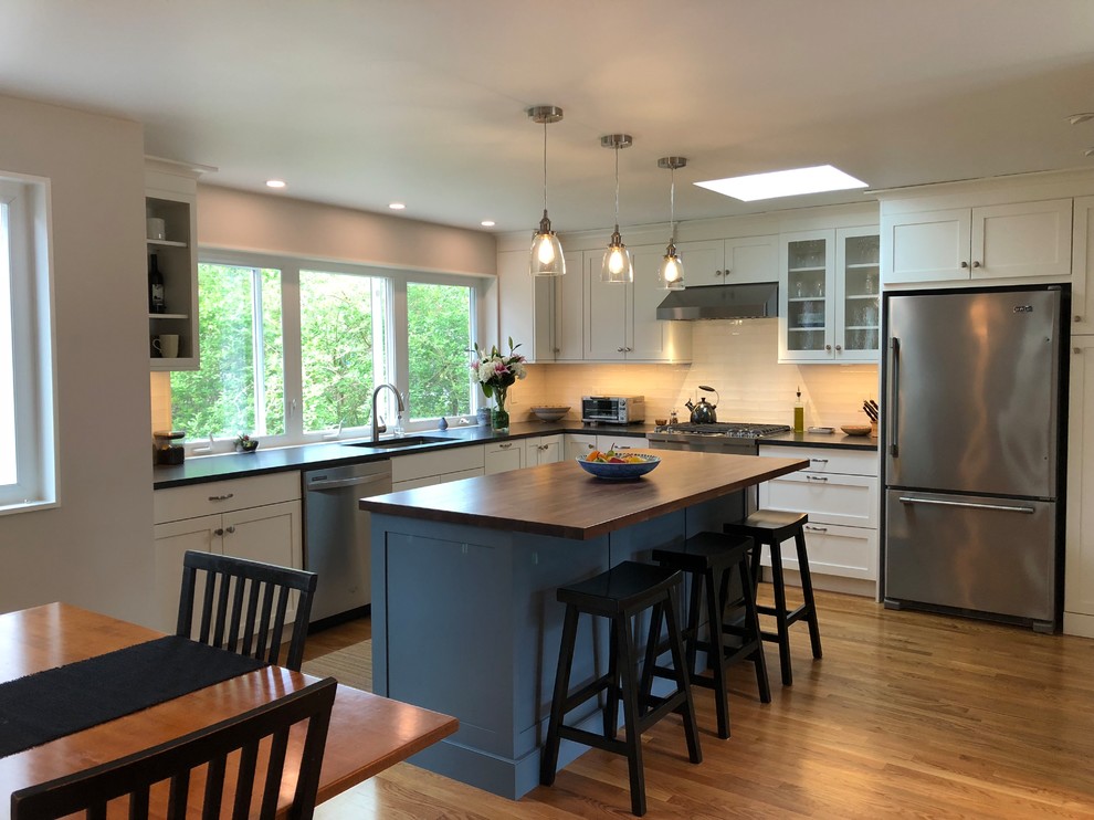 Inspiration for a mid-sized transitional l-shaped medium tone wood floor and brown floor open concept kitchen remodel in Boston with a single-bowl sink, shaker cabinets, white cabinets, granite countertops, white backsplash, window backsplash, stainless steel appliances, an island and black countertops