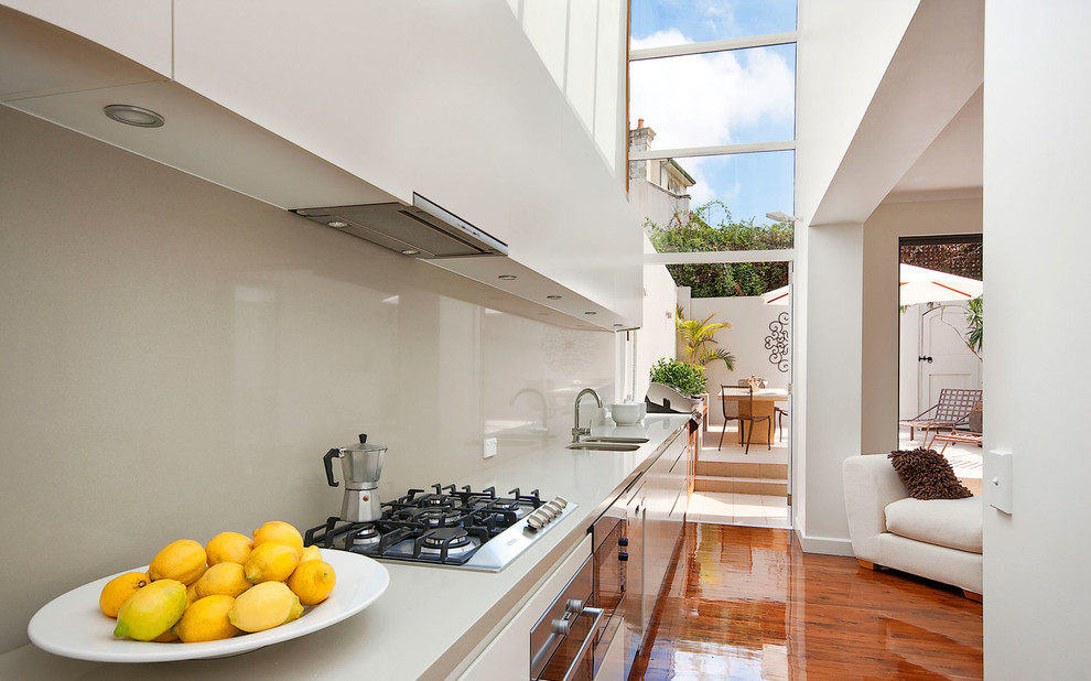 This is an example of a contemporary single-wall kitchen in Sydney with a double-bowl sink, flat-panel cabinets, white cabinets, white splashback, stainless steel appliances, medium hardwood floors and no island.