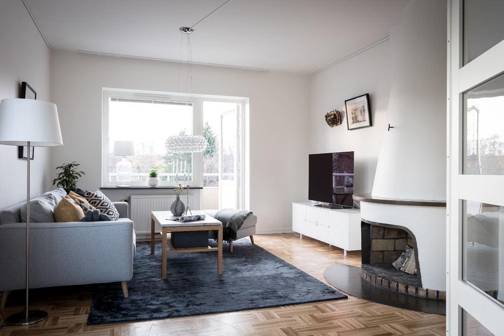 Scandinavian enclosed living room in Gothenburg with white walls, medium hardwood floors, a standard fireplace, a plaster fireplace surround, a freestanding tv and brown floor.