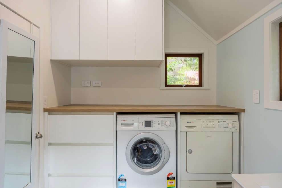 Small modern single-wall utility room in Auckland with flat-panel cabinets, white cabinets, wood benchtops, blue splashback, white walls, porcelain floors, a side-by-side washer and dryer, grey floor and brown benchtop.
