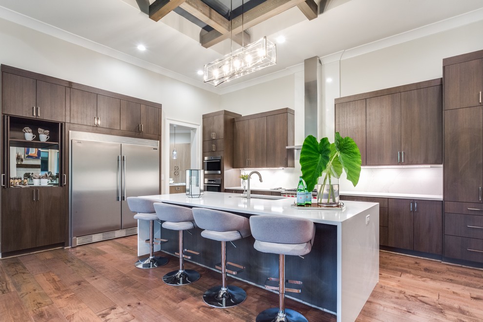 Example of a transitional medium tone wood floor and brown floor kitchen design with a single-bowl sink, flat-panel cabinets, dark wood cabinets, white backsplash, stainless steel appliances, an island and white countertops